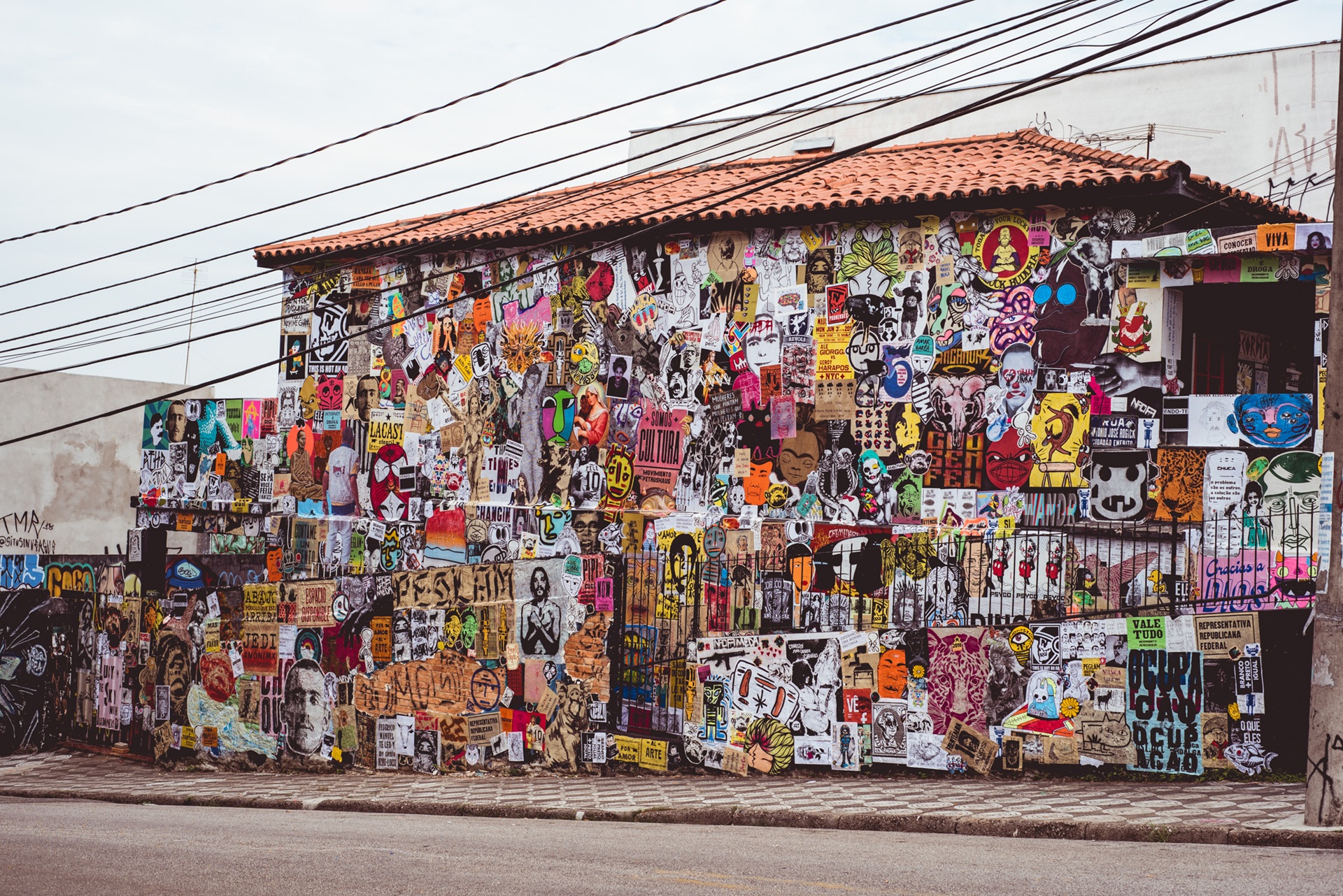 street art Fachada de casa totalmente coberta por cartazes coloridos, ilustrações e mensagens impressas, representando a técnica de lamb-lamb (cartaz colado). A composição reúne diferentes estilos e temas, criando uma colagem visual que expressa crítica social, humor e diversidade na arte urbana
