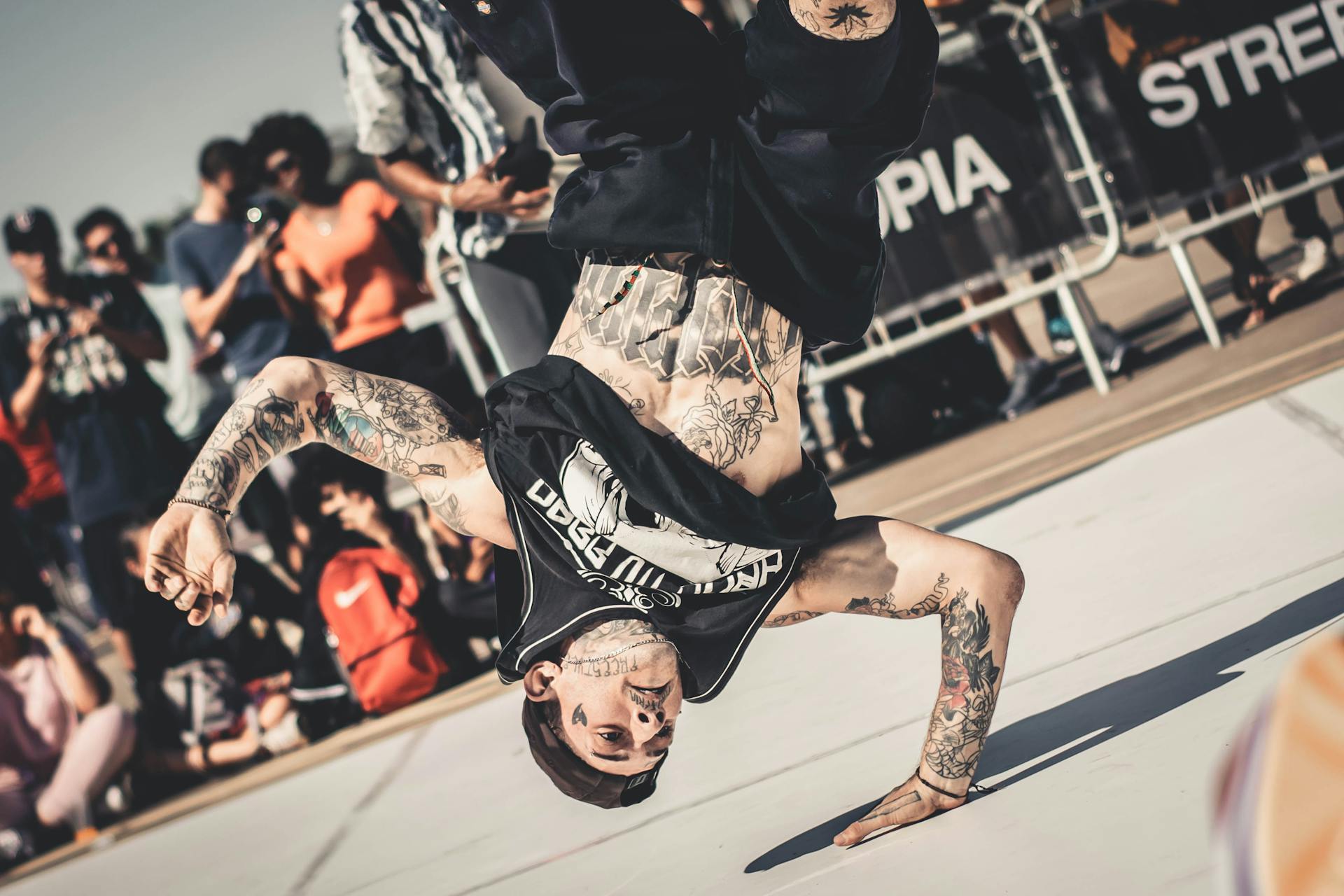 street art Dançarino executa movimento acrobático de breakdance apoiado em uma mão, durante apresentação de rua com público ao fundo