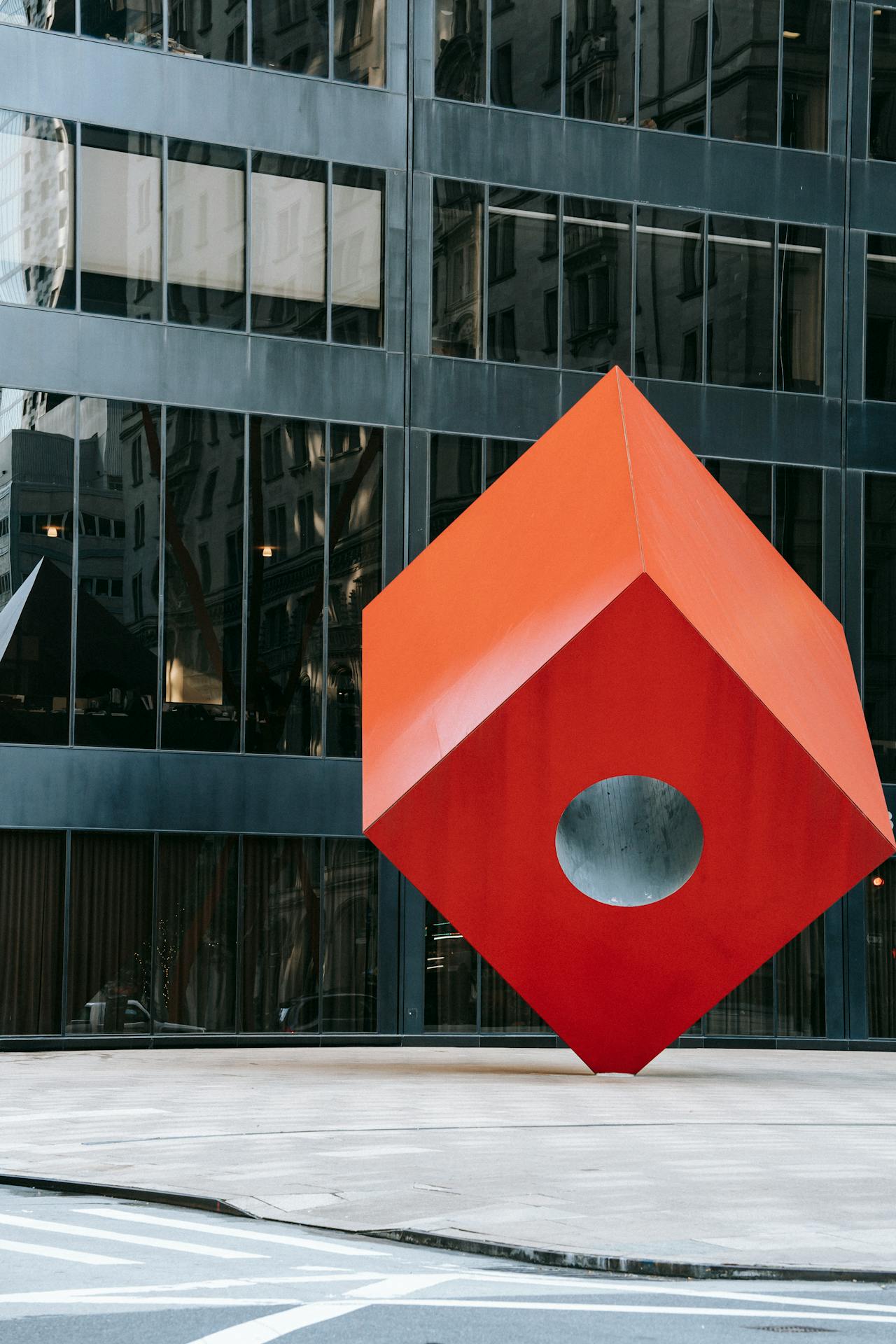 street art Grande escultura cúbica vermelha com um furo circular no centro, instalada em área urbana cercada por prédios espelhados