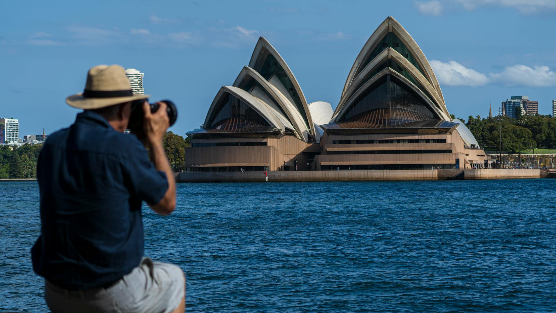 Fotógrafo de chapéu captura a Sydney Opera House à beira do porto, com as suas icônicas velas ao fundo
