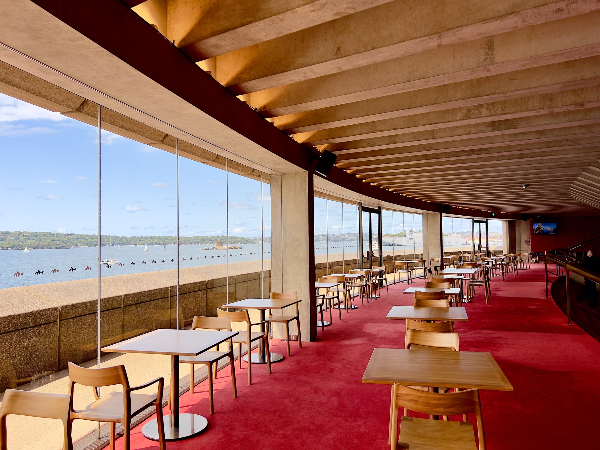 Área de convivência do Joan Sutherland Theatre, na Sydney Opera House, com mesas e cadeiras dispostas sobre carpete vermelho e janelas de vidro do piso ao teto, revelando a paisagem do porto ao fundo