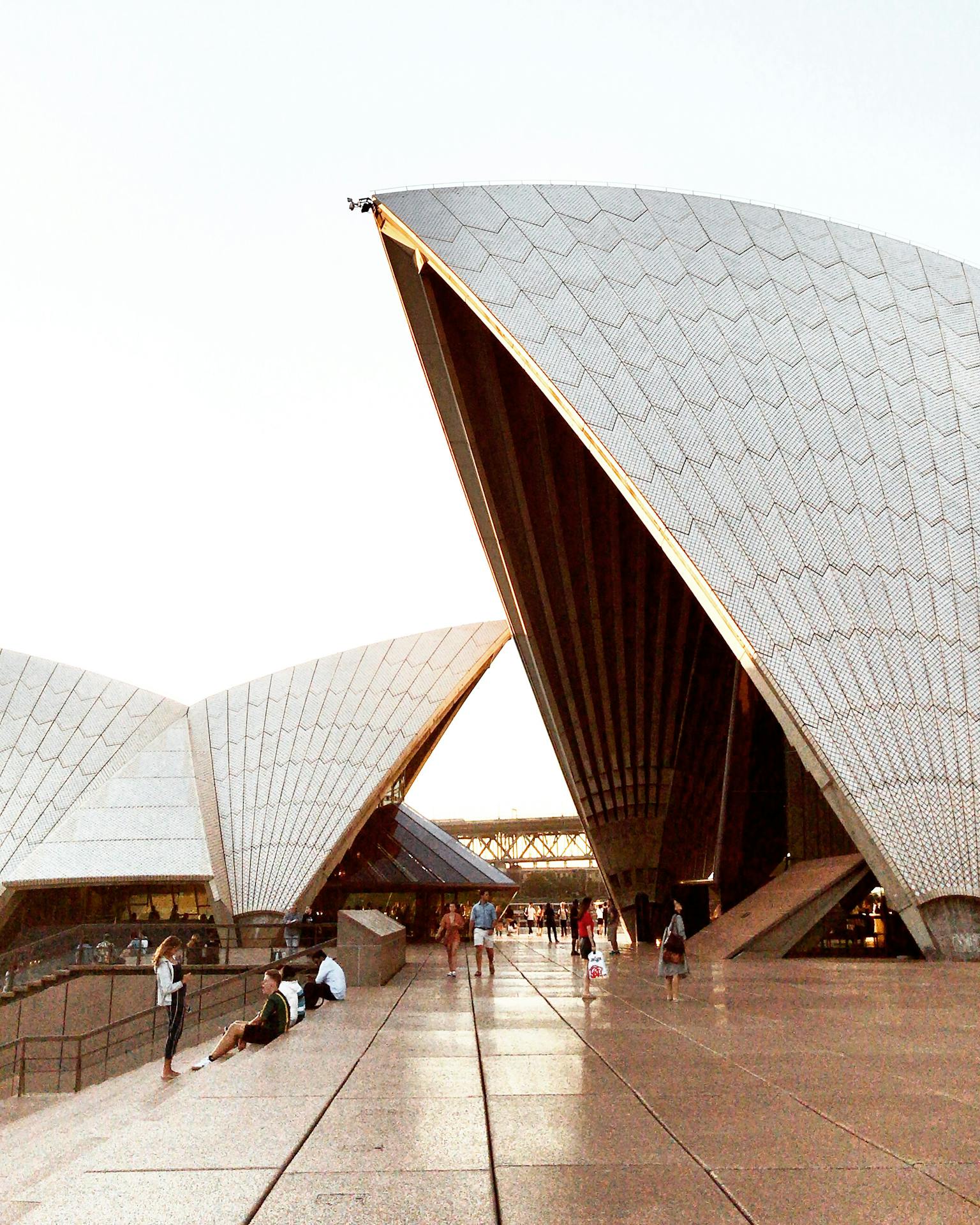 Vista externa da Sydney Opera House ao entardecer, com pessoas caminhando pelo espaço