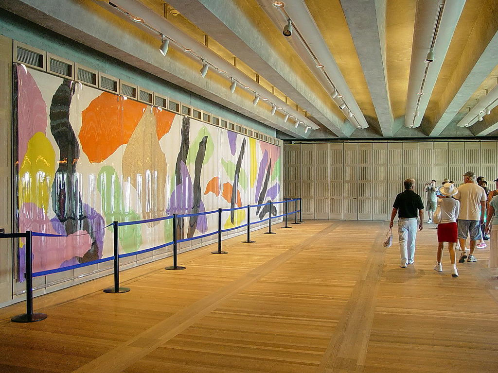 Interior da Utzon Room, na Sydney Opera House, com piso de madeira e teto com vigas aparentes. A grande tapeçaria colorida assinada por Jørn Utzon ocupa uma das paredes, enquanto visitantes exploram o ambiente cultural e acolhedor