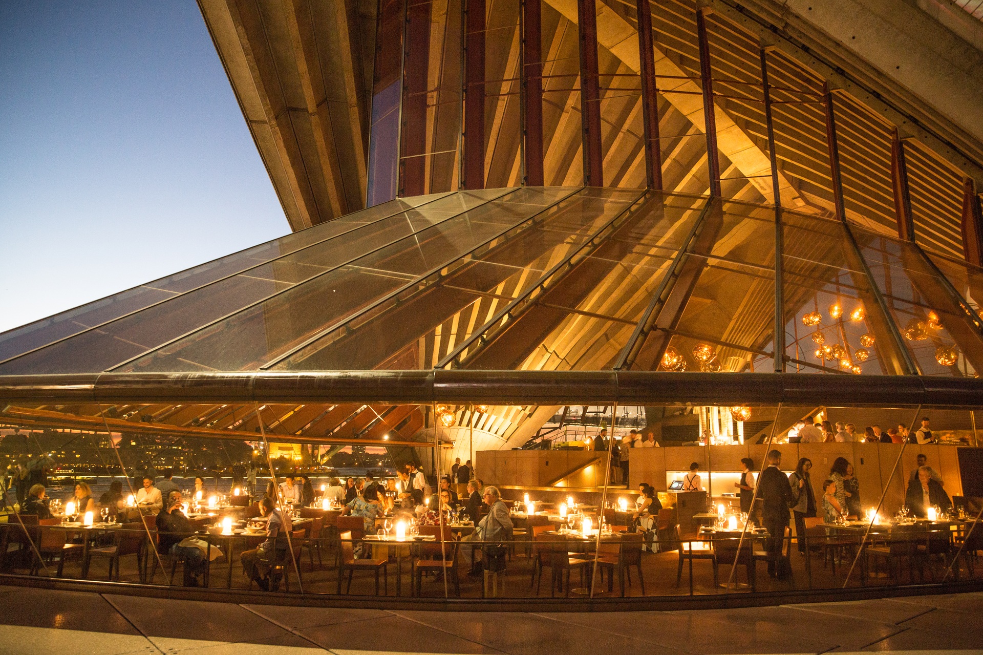 Vista externa de restaurante nas Yallamundi Rooms, na Sydney Opera House, com janelas de vidro que revelam pessoas reunidas em mesas iluminadas
