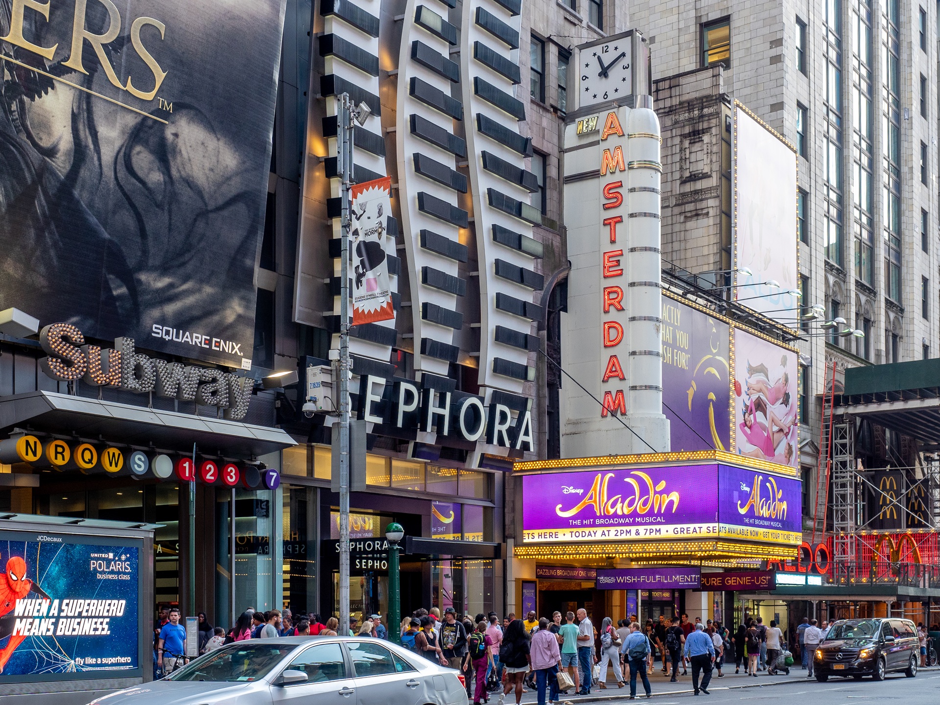 Fachada do New Amsterdam Theatre na Broadway, em Nova York, com letreiros iluminados do musical Aladdin e movimento intenso de pedestres e carros na rua
