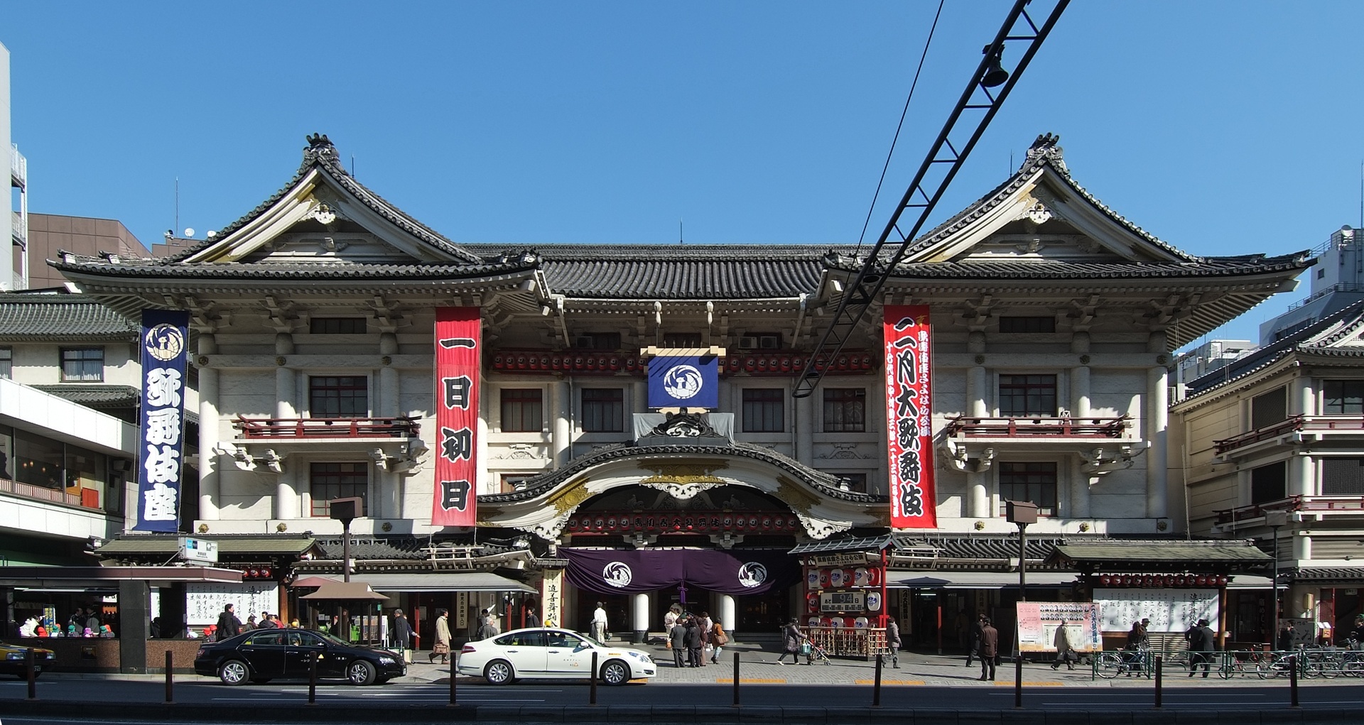 Fachada do Kabuki-za, em Tóquio, Japão, com telhados característicos, ornamentos detalhados e bandeiras verticais vermelhas e azuis, em uma rua movimentada com pedestres e carros