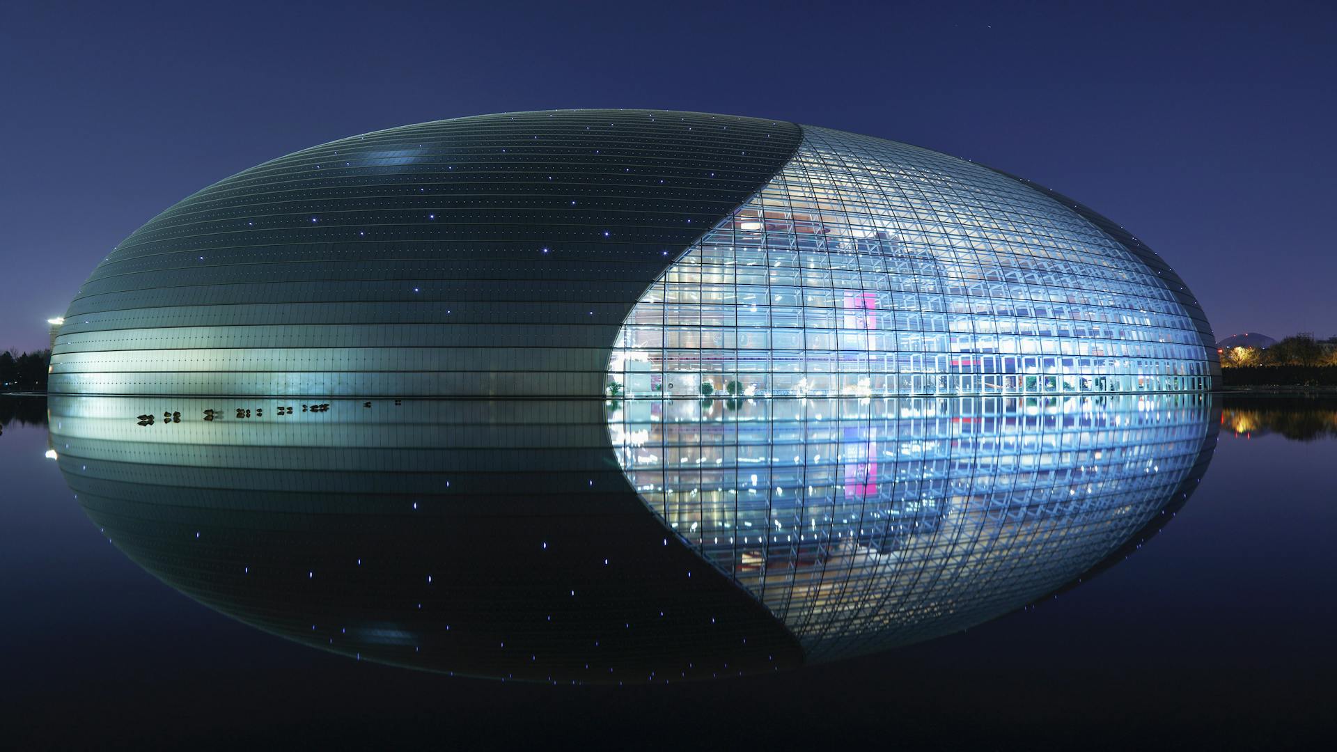 Vista noturna do National Centre for the Performing Arts of China, em Pequim, com estrutura oval de vidro e titânio refletida na água ao redor, iluminada por luzes internas