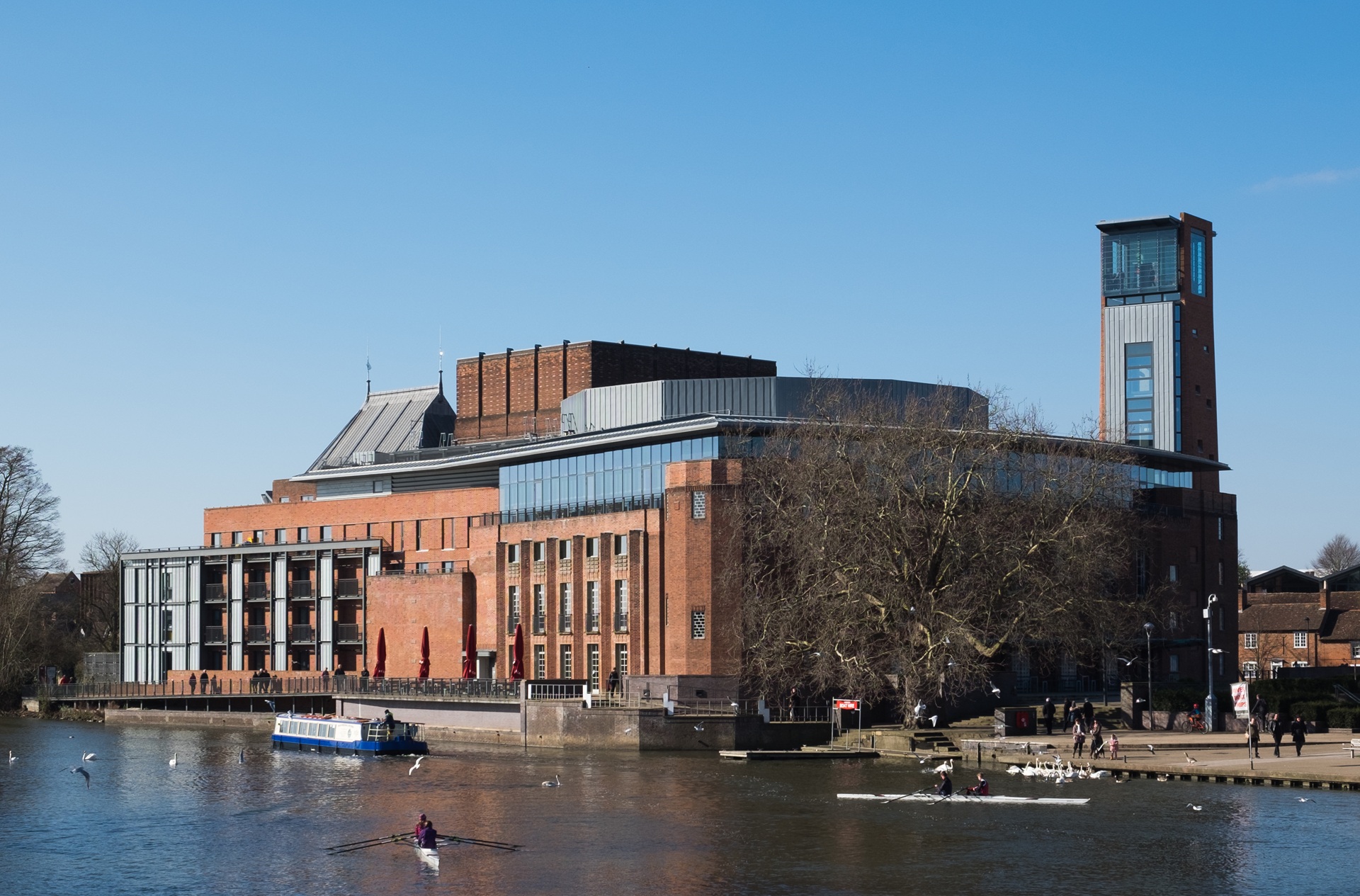 Vista do Royal Shakespeare Theatre, em Stratford-upon-Avon, Inglaterra, com fachada de tijolos vermelhos e torre de observação, localizado à beira do rio Avon, em dia de céu azul