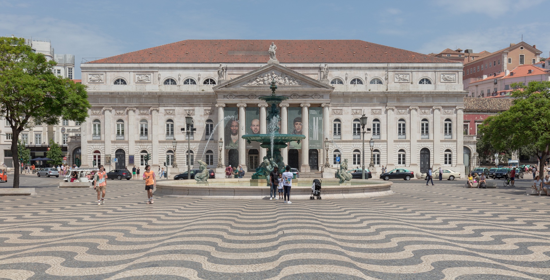 Vista frontal do Teatro Nacional D. Maria II, em Lisboa, Portugal, com fachada neoclássica e fonte em primeiro plano na Praça do Rossio, em um dia ensolarado
