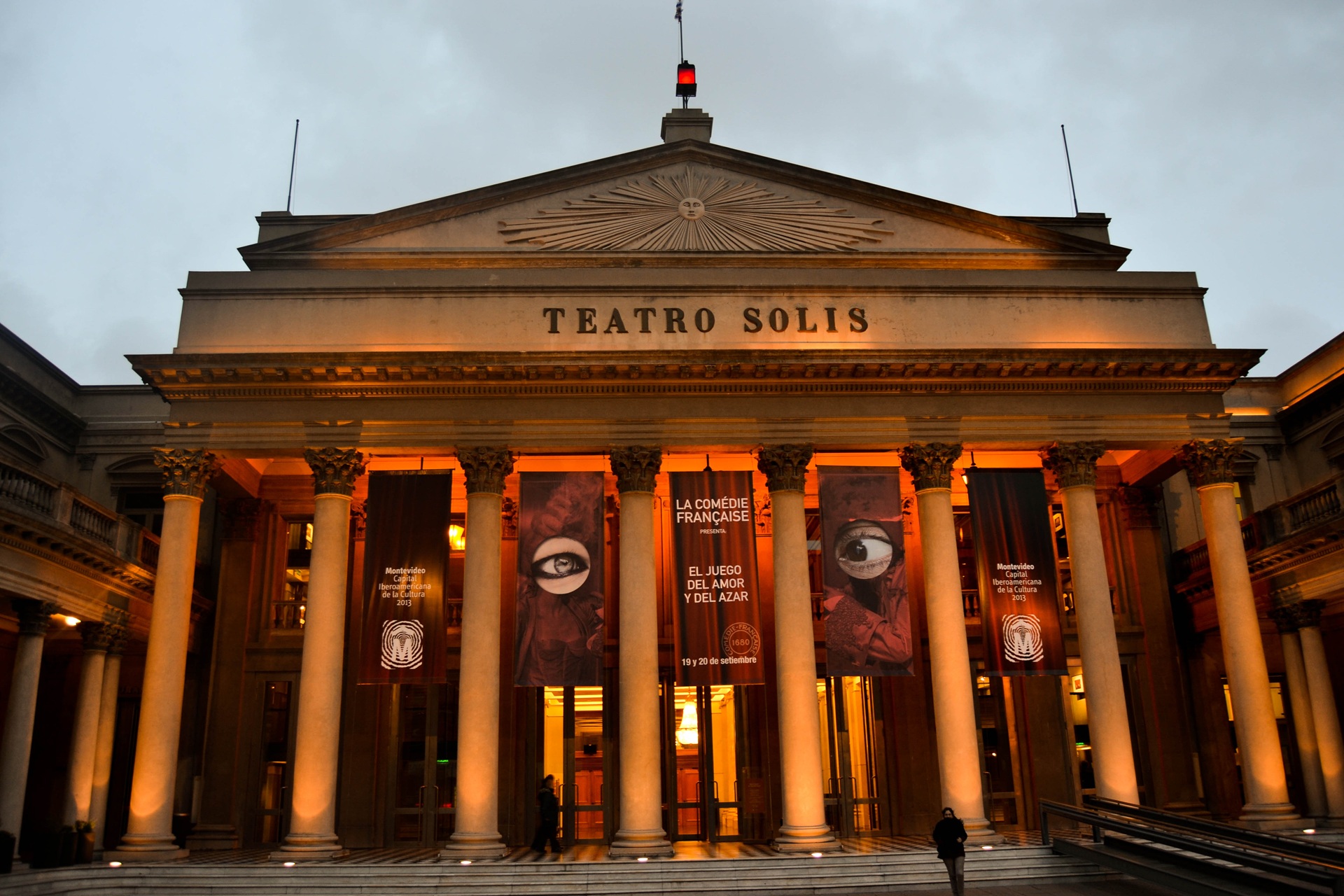 Fachada iluminada do Teatro Solís, em Montevidéu, Uruguai, com colunas clássicas e banners de divulgação pendurados na entrada, durante o anoitecer
