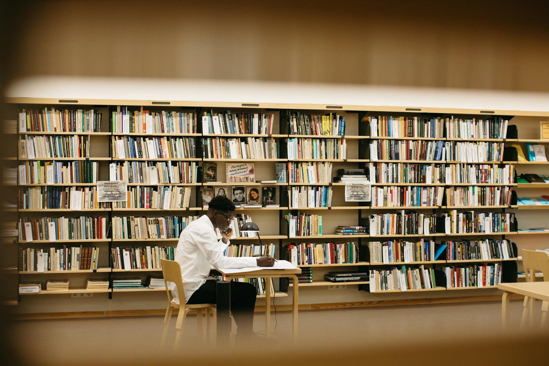 Pessoa sentada à mesa em uma biblioteca iluminada, concentrada em anotações, com estantes cheias de livros ao fundo