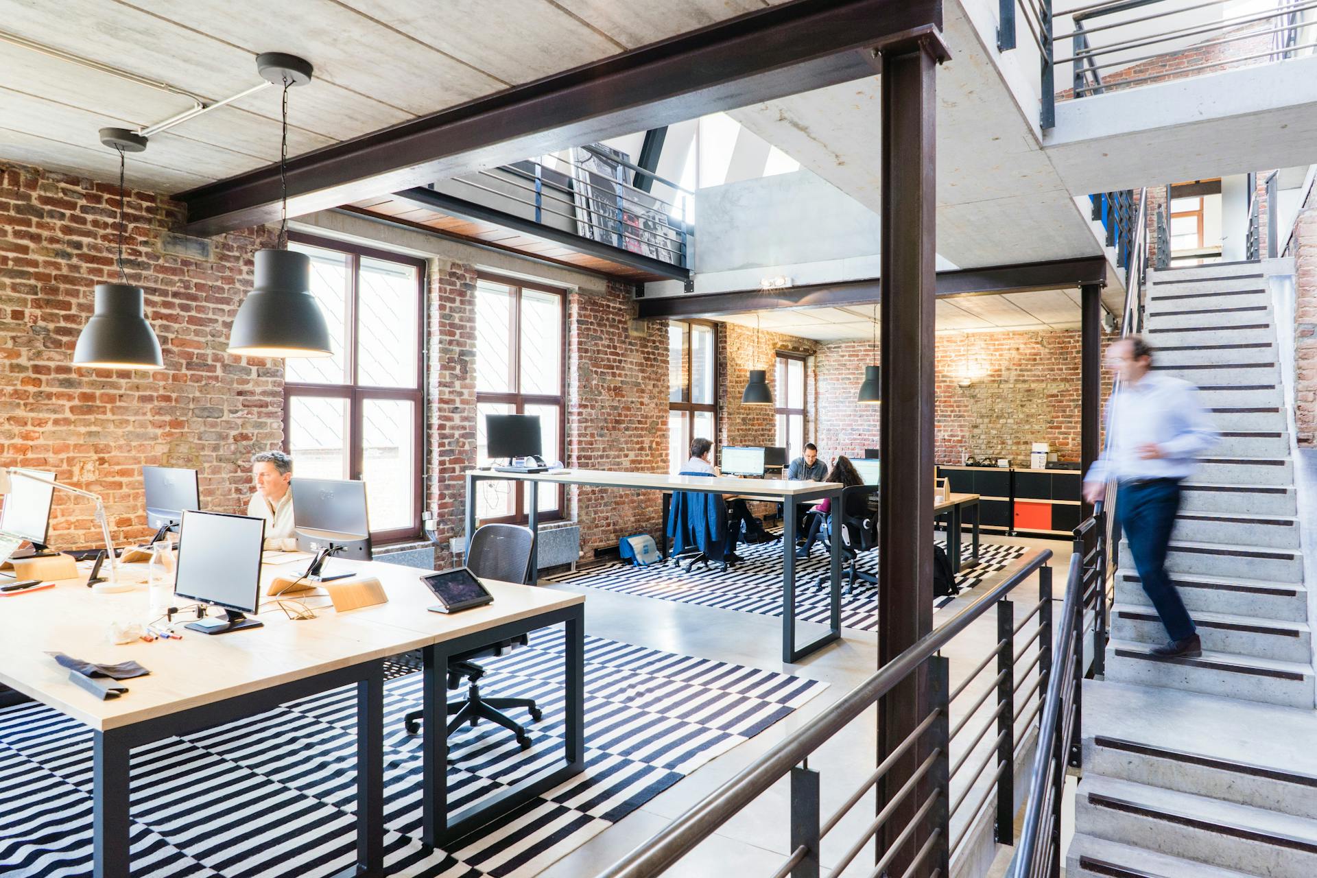 Espaço de coworking com estrutura moderna, paredes de tijolos aparentes e janelas amplas; pessoas trabalham em mesas coletivas sob iluminação natural