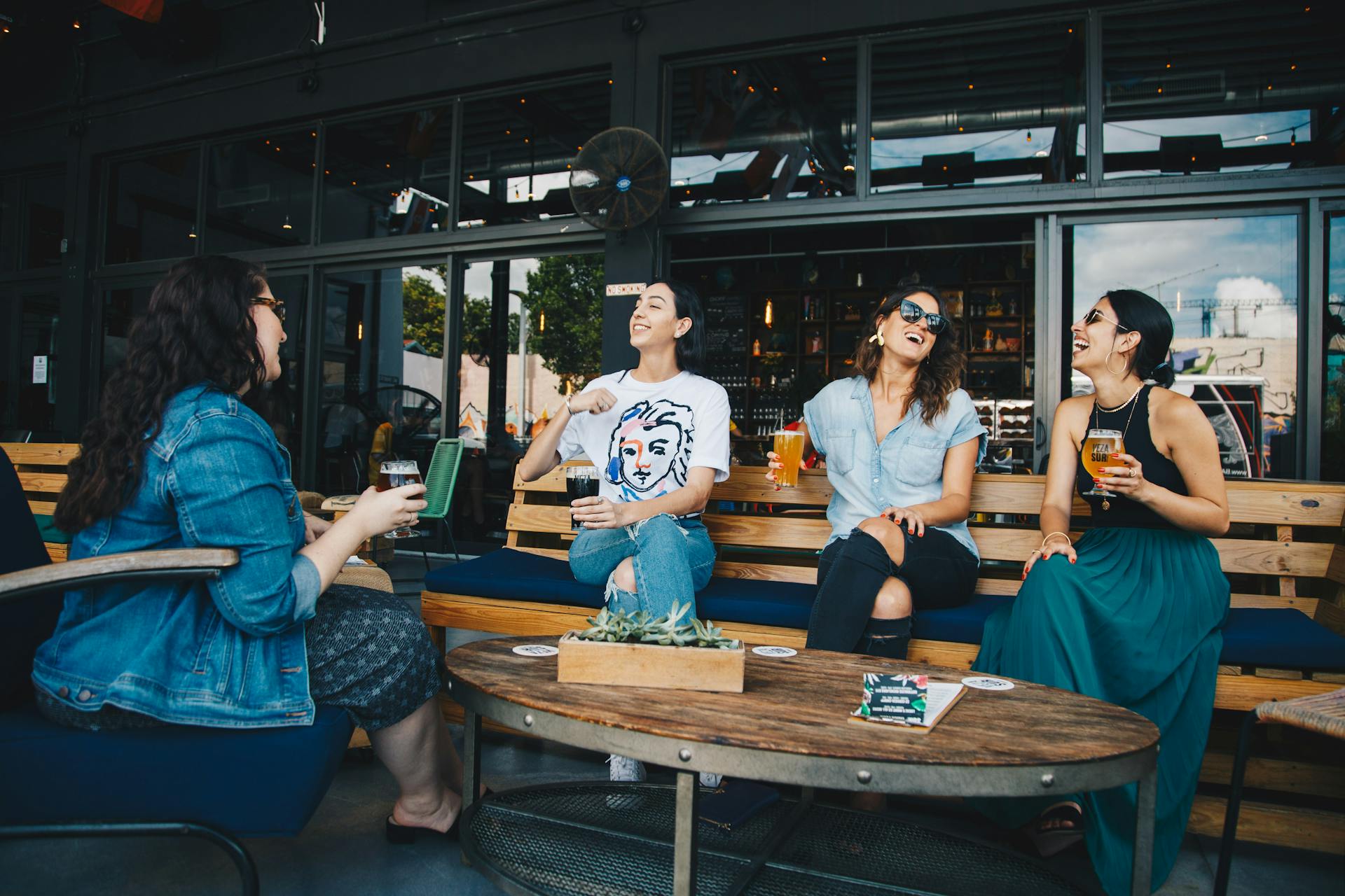 Quatro pessoas sentadas em um ambiente externo de bar ou cafeteria, sorrindo e conversando enquanto seguram copos de bebida