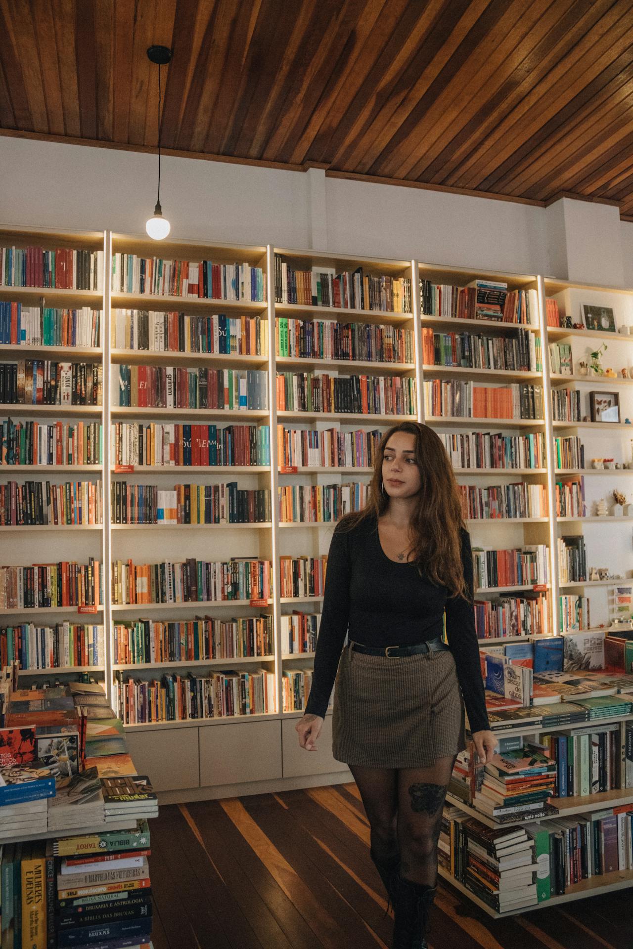 Mulher caminha entre estantes repletas de livros em uma livraria iluminada por luz suave e aconchegante