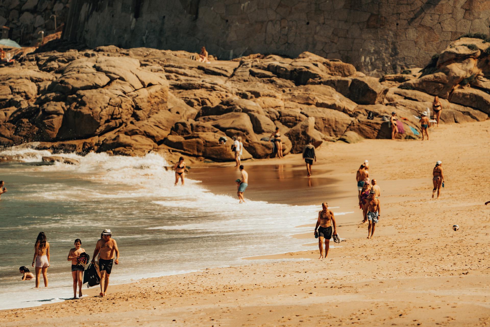 Cena de praia com pessoas caminhando pela areia, entrando no mar e descansando próximas às rochas