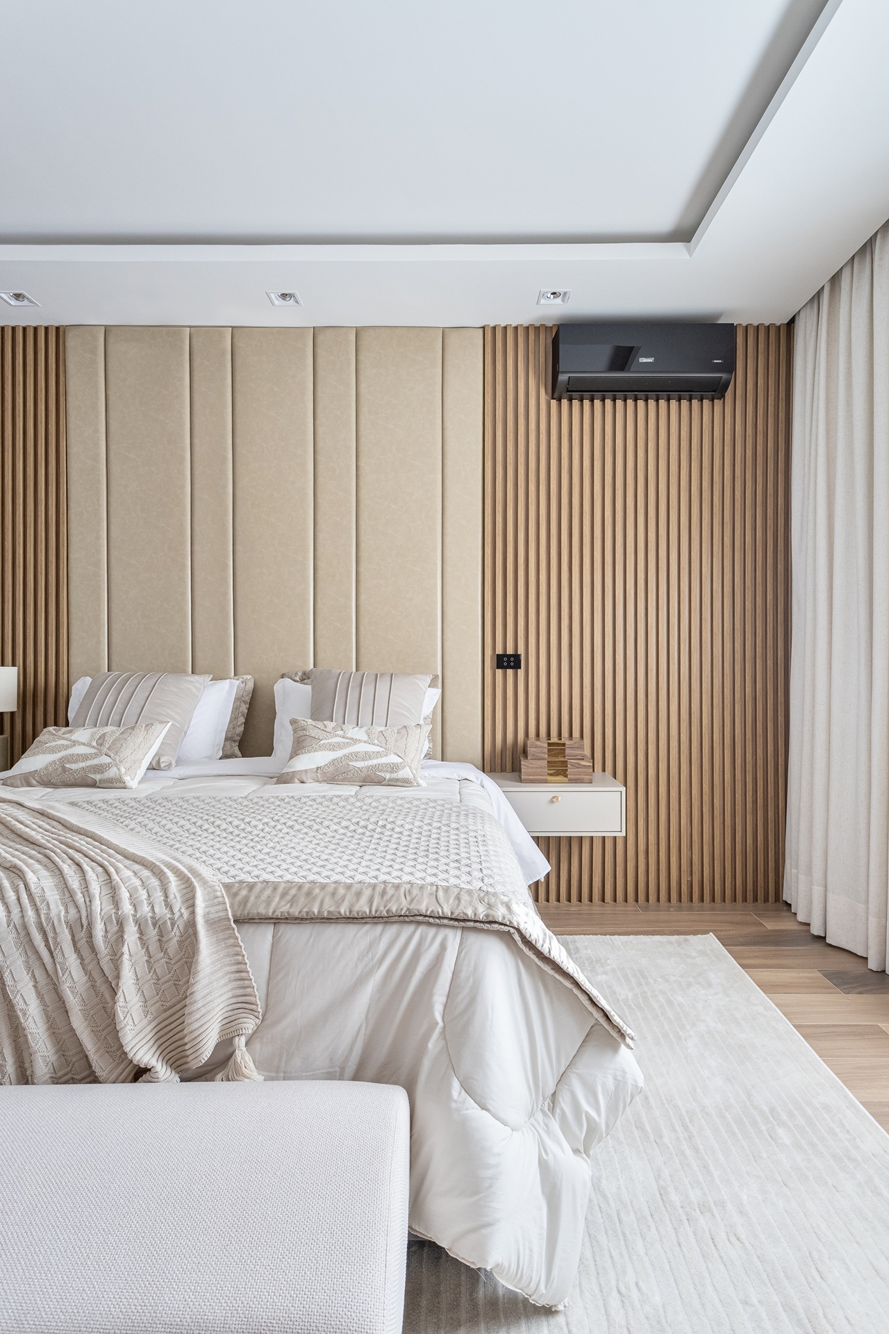Quarto com cama de casal com roupas de cama beges, piso amadeirado e painel ripado atrás da cama com cabeceira estofada
