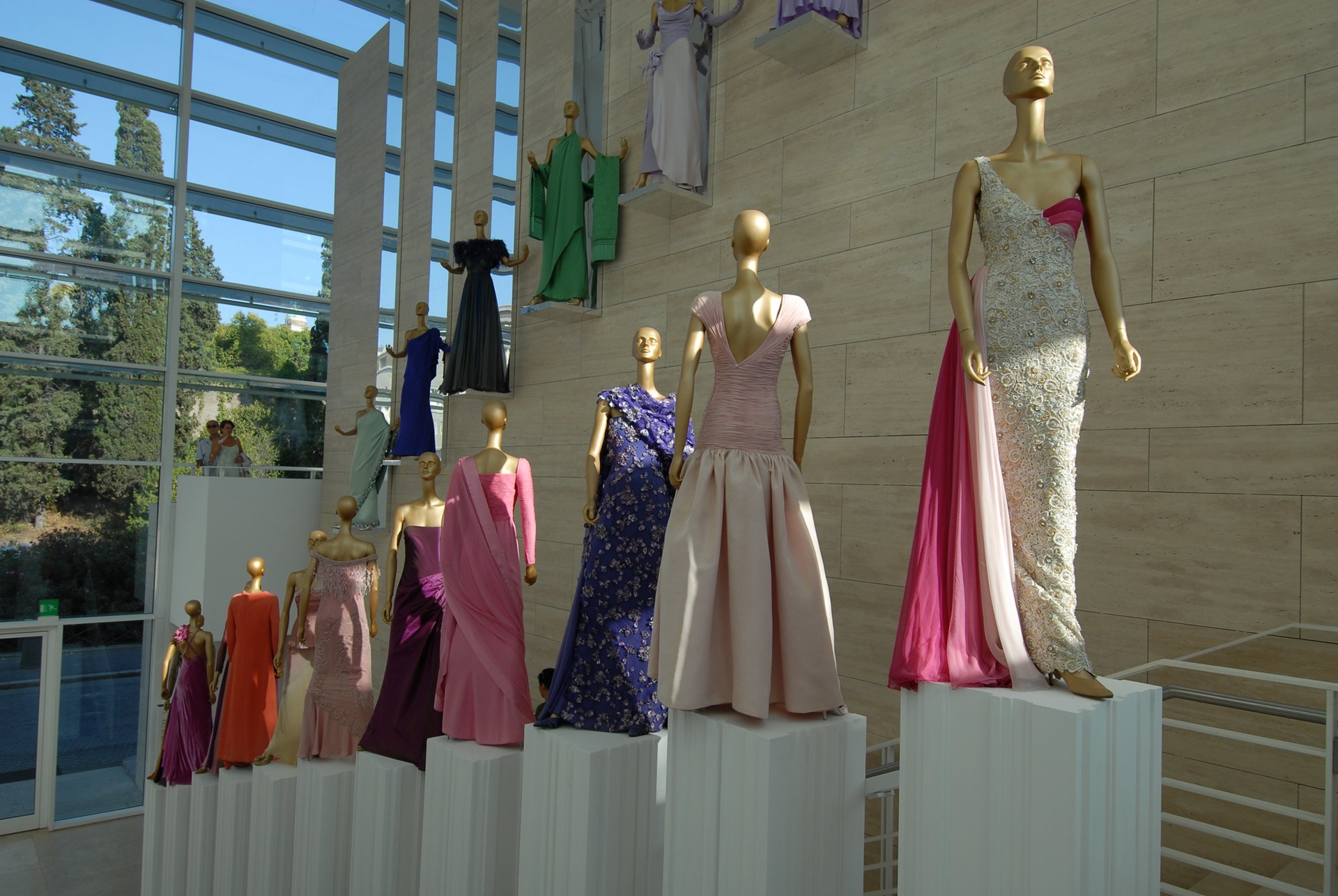 Exposição em um museu na Itália com manequins vestidos em criações da marca Valentino; os modelos exibem vestidos de alta-costura em tons de rosa, roxo, azul e verde, dispostos em pedestais brancos sob luz natural que entra por amplas janelas de vidro
