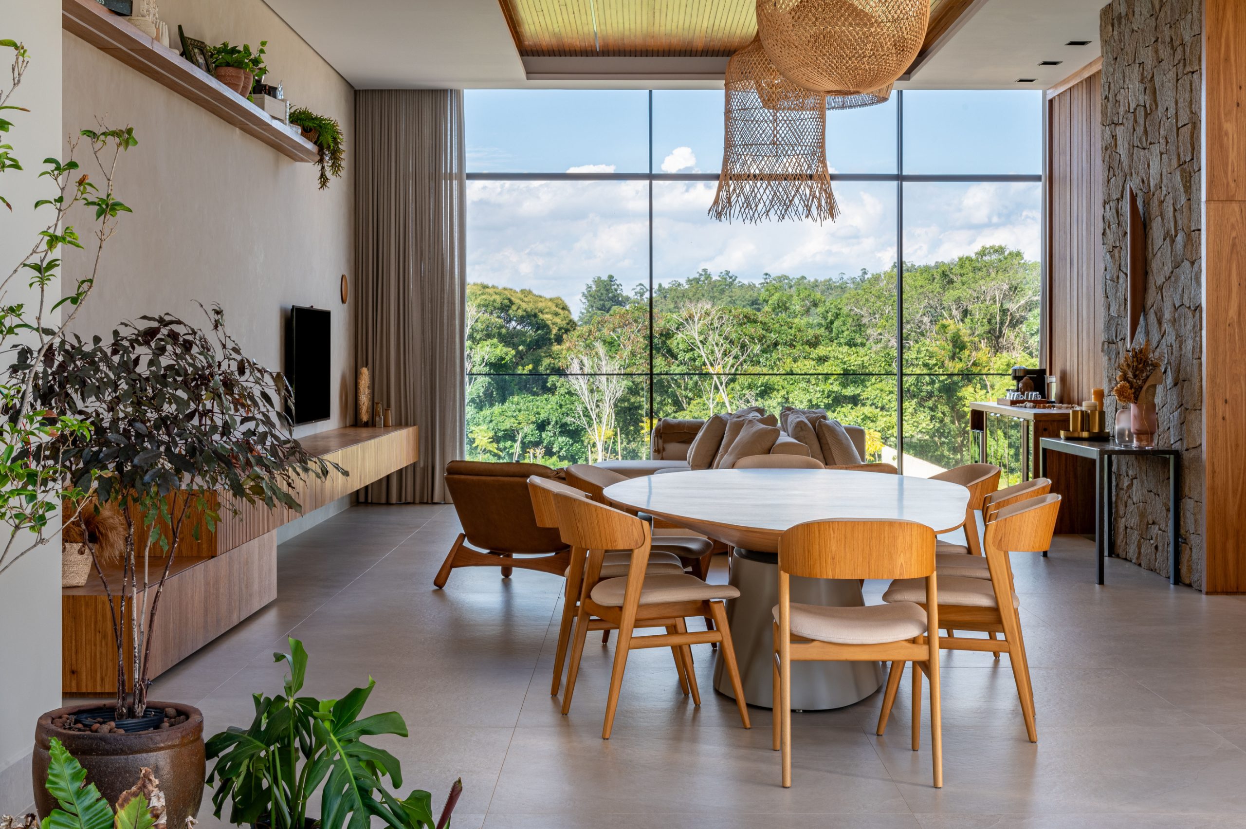 Living com sofá, televisão e mesa de jantar em formato orgânico com ampla parede de vidro com vista para área verde
