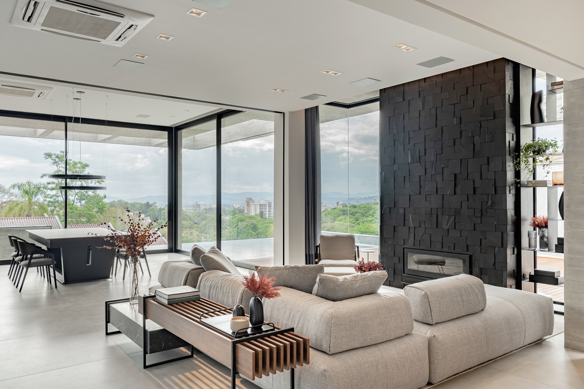 Sala de estar em tons claros com parede com lareira com revestimento escuro e fechamento em vidro com vista para o horizonte da cidade