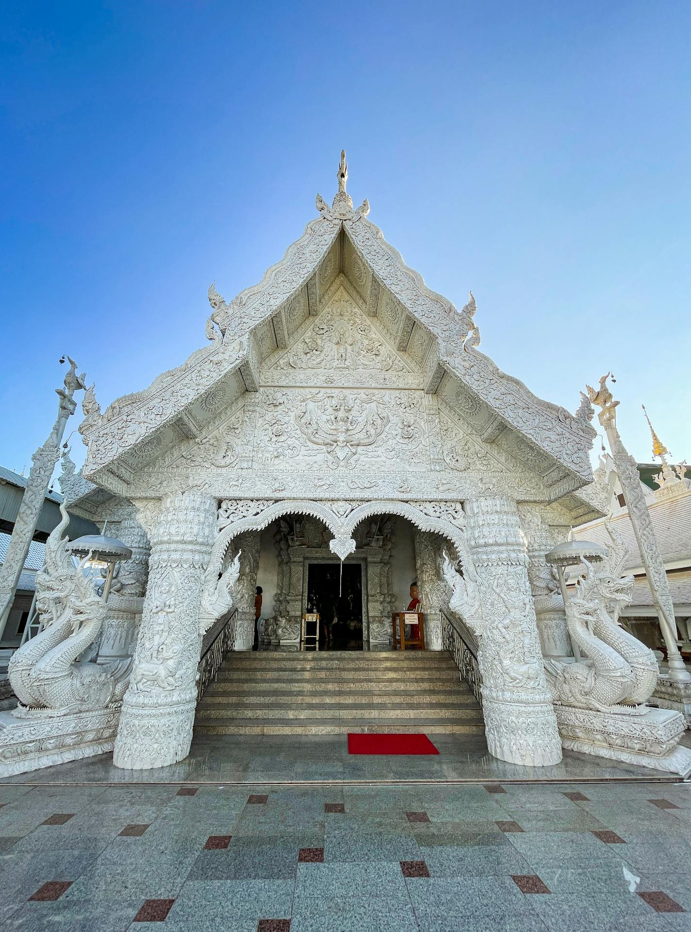 Fachada frontal do ubosot do Wat Rong Khun, o Templo Branco de Chiang Rai, com escadaria central e colunas decoradas por serpentes mitológicas Naga