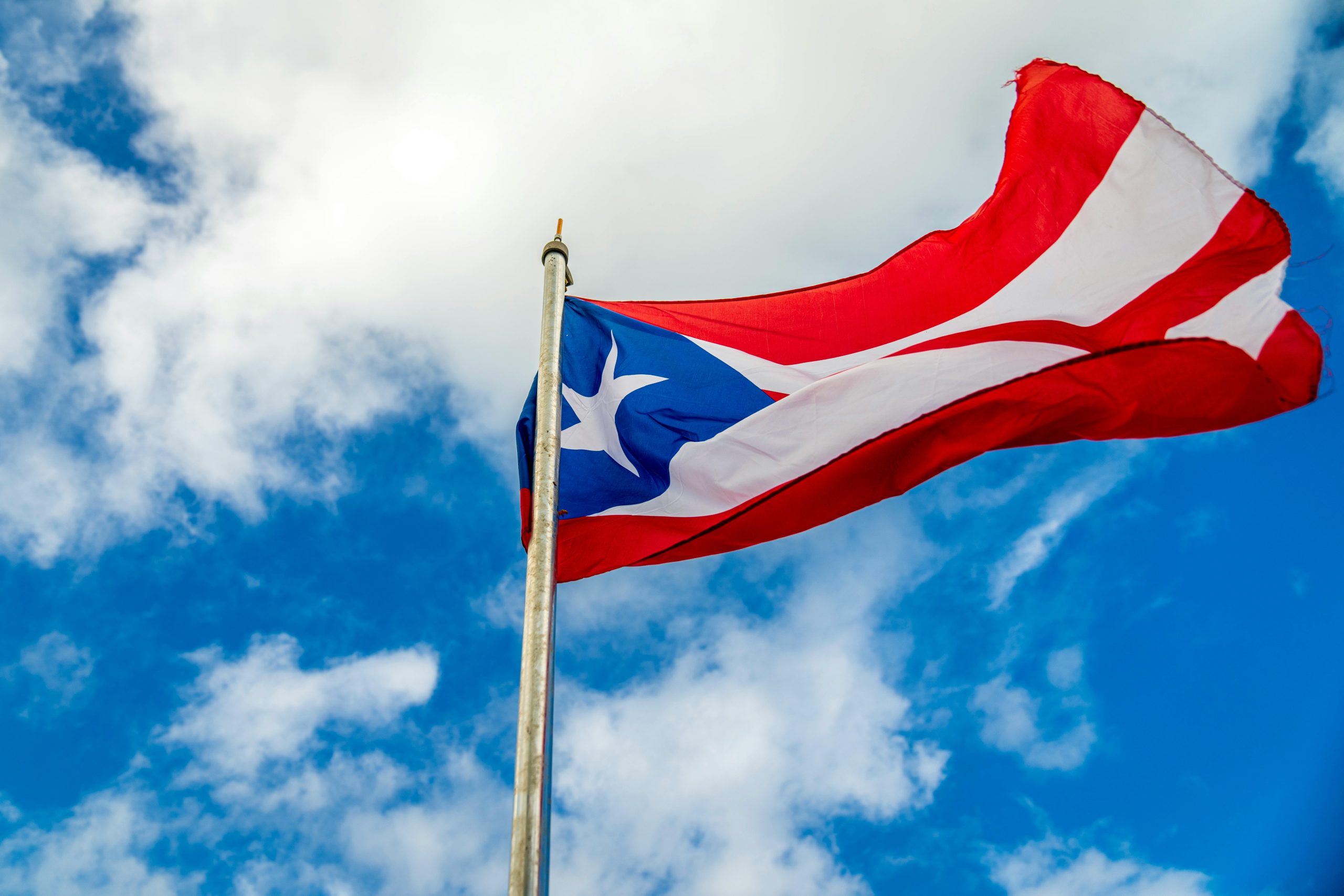 Bandeira de Porto Rico tremulando em um mastro metálico, com triângulo azul e estrela branca à esquerda e faixas vermelhas e brancas horizontais, contra um céu azul com nuvens brancas