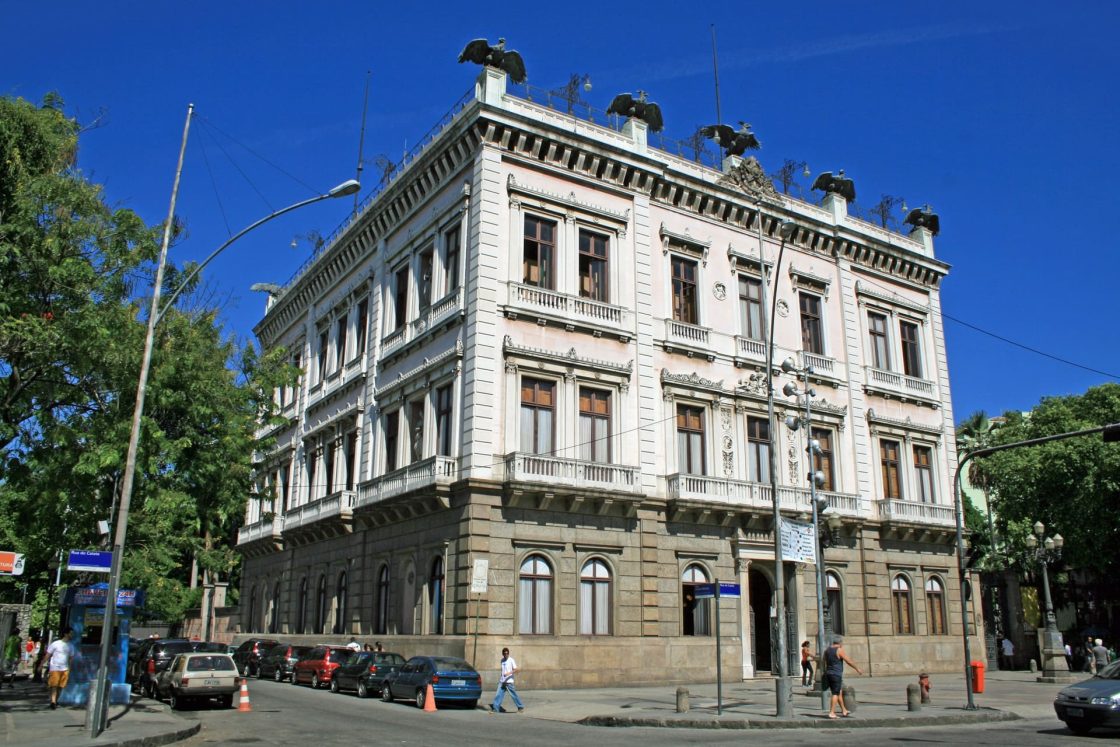 Imagem externa do Palácio do Catete mostrando o prédio histórico com detalhes ornamentais, situado em esquina arborizada da cidade