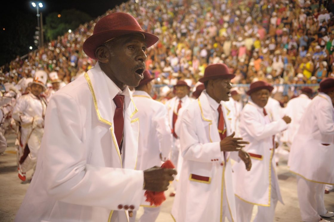 Integrantes de escola de samba vestidos de branco com chapéu e gravata desfilam no sambódromo, representando a figura tradicional do malandro durante apresentação carnavalesca