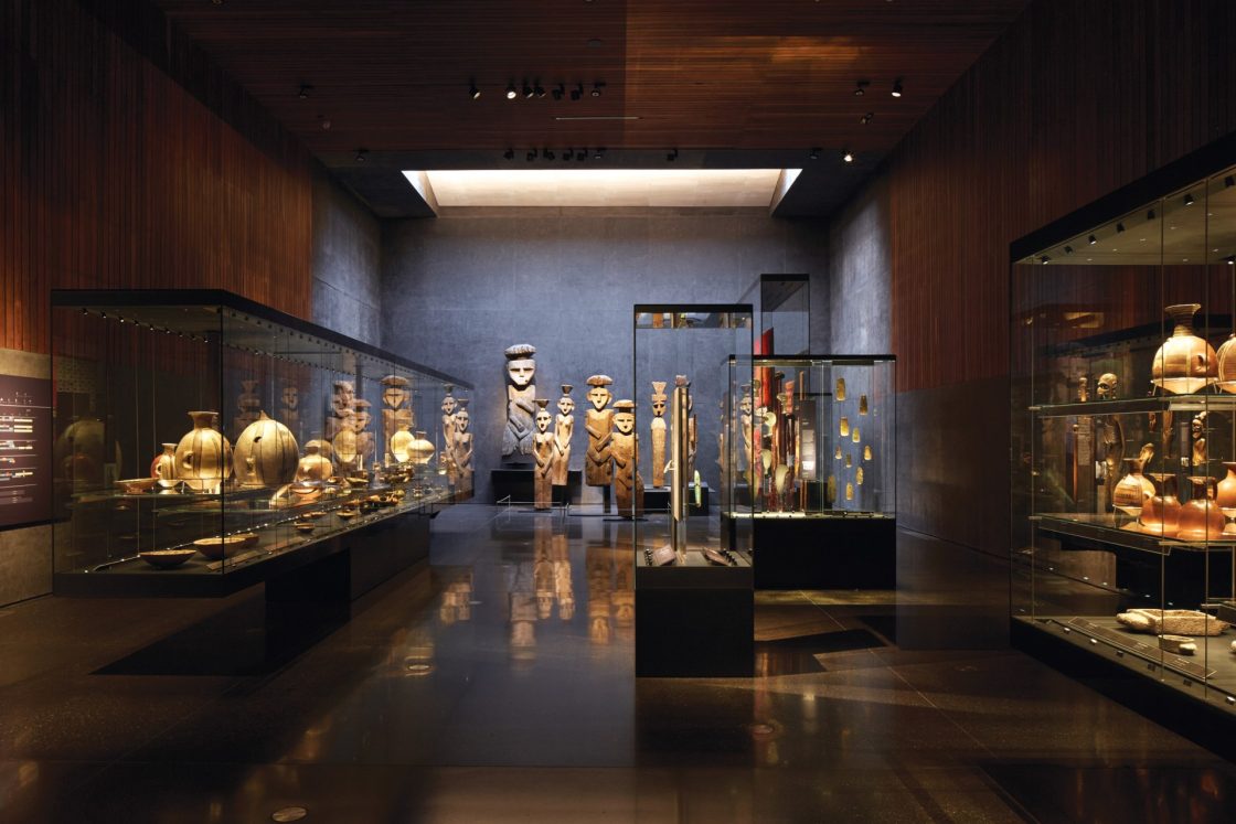 Interior de museu com luz baixa e artefatos como esculturas e vasos expostos em vitrines