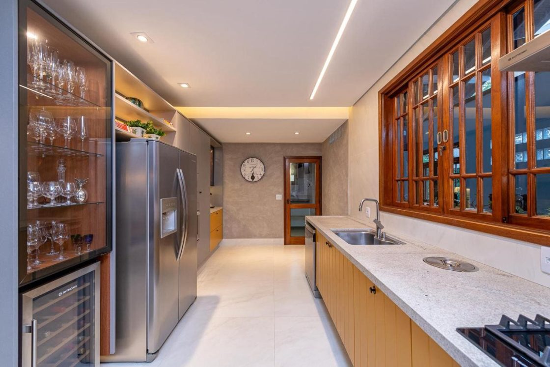 Cozinha reformada com proposta minimalista e inspiração colonial, piso e parede revestidos com o mesmo porcelanato claro, esquadrias e portas em madeira, mobiliário em tom amadeirado e eletrodomésticos em inox compondo ambiente harmonioso