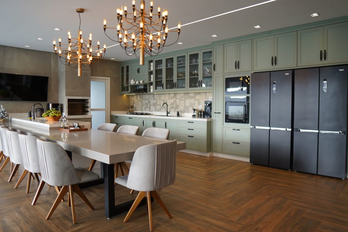 Sala de jantar integrada à cozinha com piso em porcelanato amadeirado, marcenaria verde, mesa ampla com cadeiras estofadas e iluminação pendente contemporânea