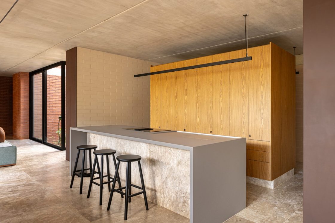 Cozinha com piso rústico, ilha com acabamento cinza, banquetas pretas, armário de madeira ao fundo e parede com revestimento de tijolinho off white ao lado