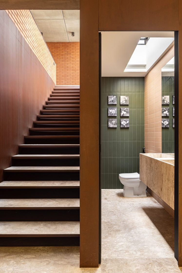 Escada com parede em aço corten e, ao lado, banheiro com revestimentos de parede verde e bege, em formato retangular, e piso neutro e rústico