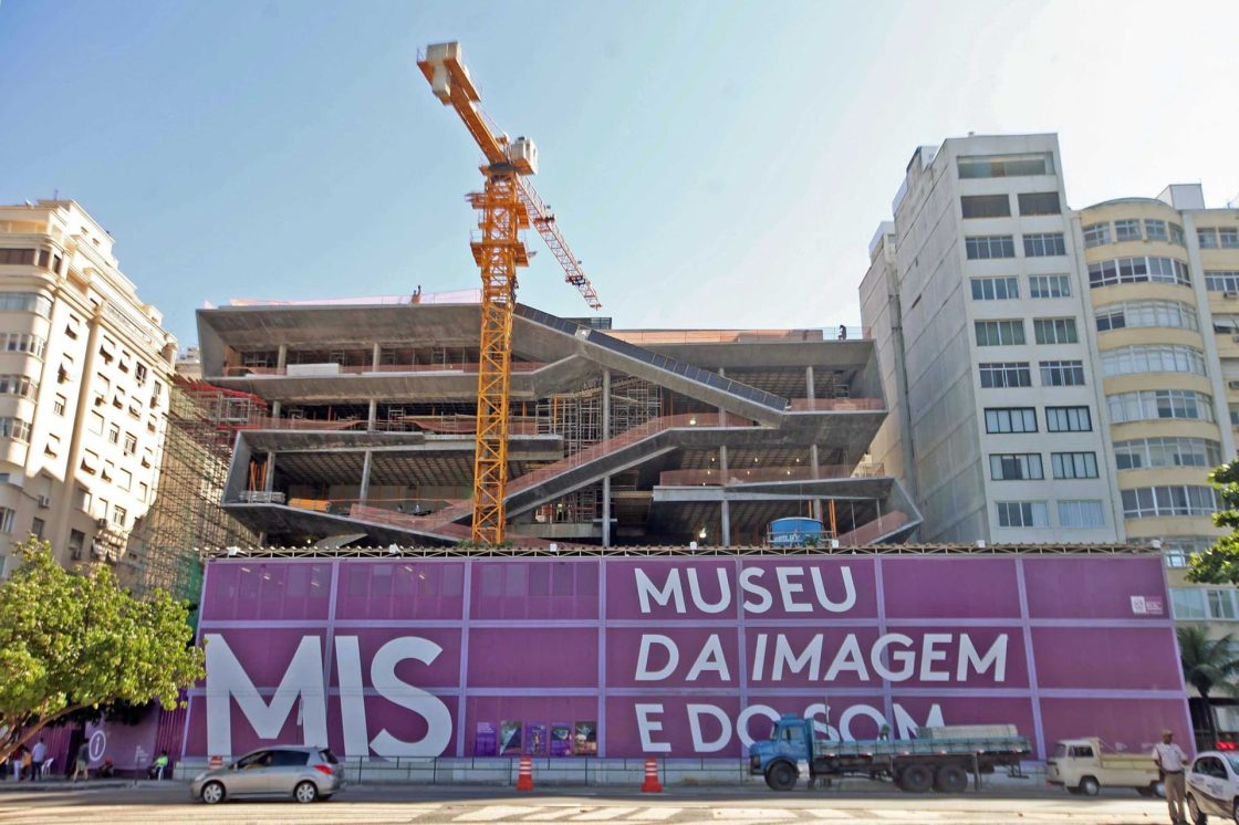 Museu da Imagem e do Som, no Rio de Janeiro, em construção, com estrutura de concreto em níveis inclinados, guindaste central e tapume com identificação do projeto na fachada