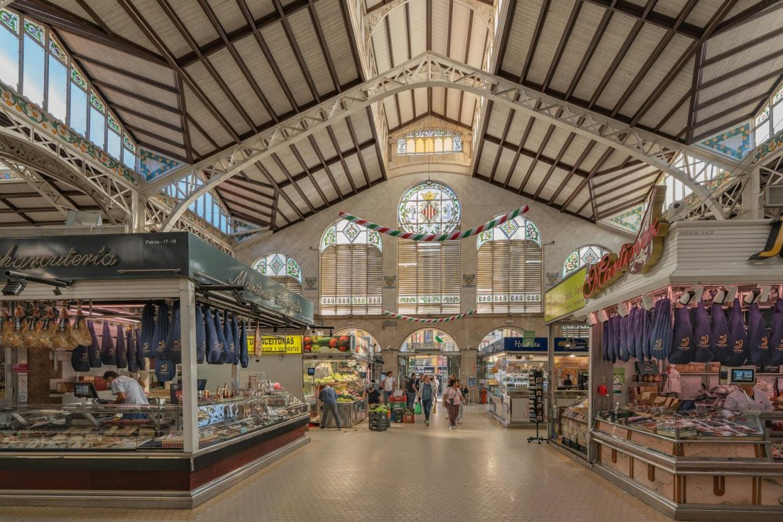 Interior do Mercado Central de Valência, com cobertura metálica arqueada, vitrais decorativos, corredores amplos e bancas de carnes, embutidos e frutas organizadas sob luz natural que atravessa as janelas ornamentadas