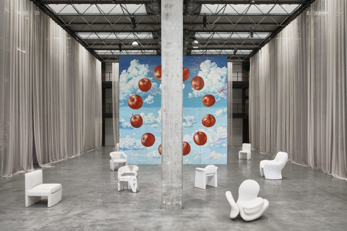 Em ambiente industrial em tons acinzentados, poltronas brancas aparecem dispostas uma longe da outra, com painel ao fundo que tem céu com nuvens e círculo grande formado por maçãs