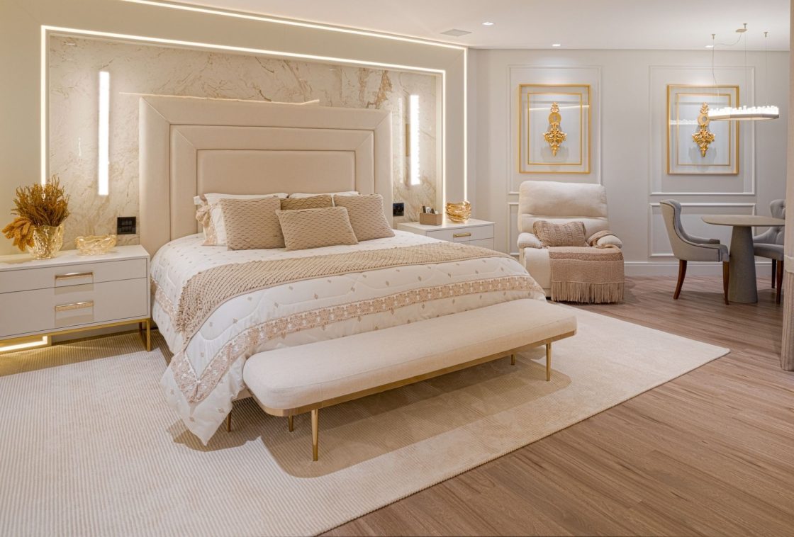 Dormitório luxuoso, com cama imponente em tons de bege, apresentando cabeceira trabalhada, enxoval matelassado e iluminação indireta