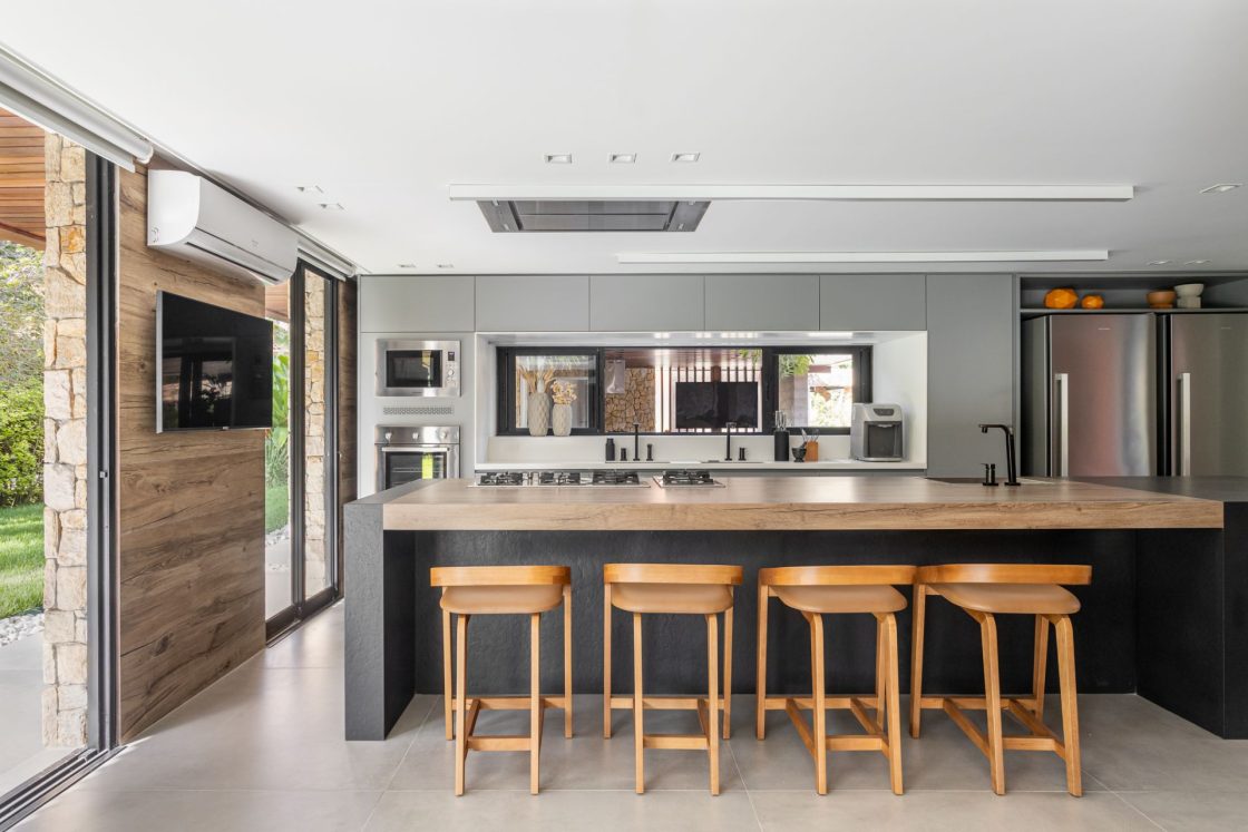 Cozinha com ilha com revestimento preto e balcão amadeirado, com banquetas em madeira e revestimento claro no piso