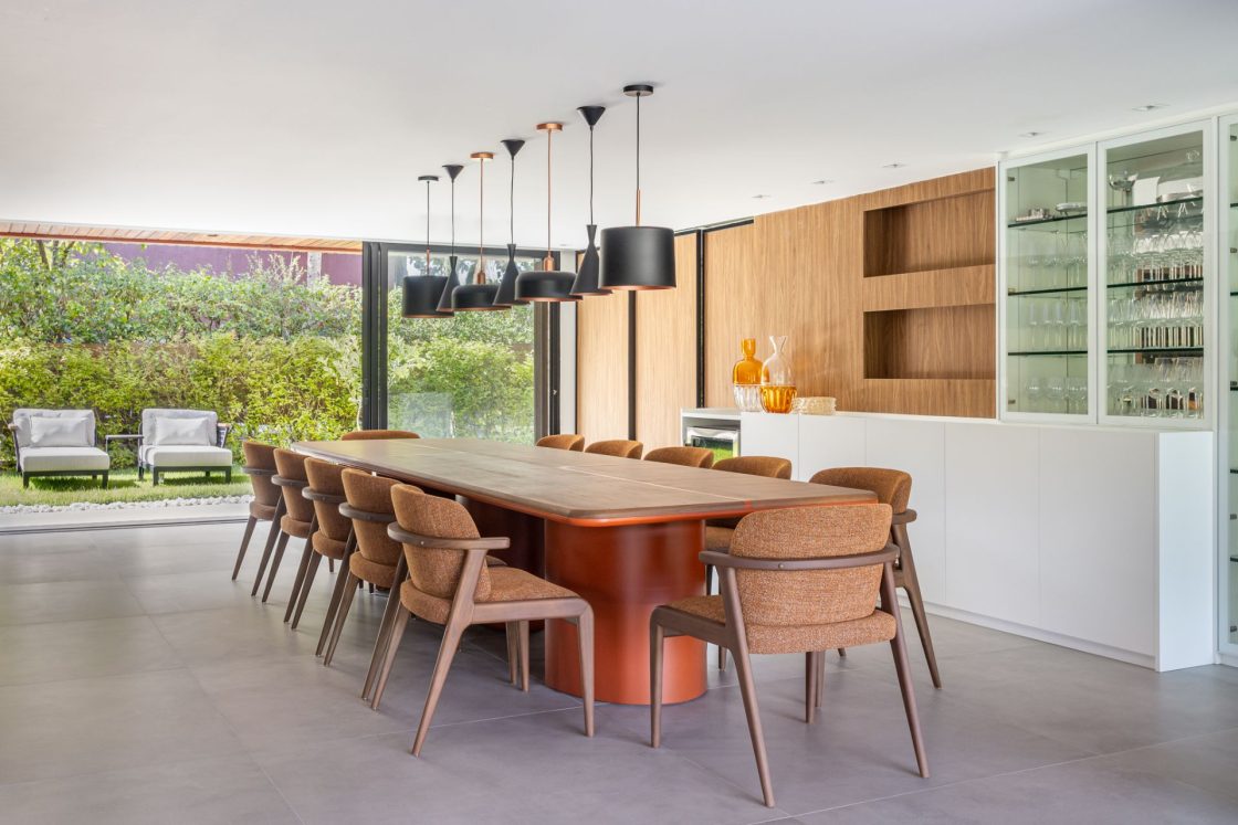 Sala de jantar ampla com piso claro, parede com revestimento amadeirado, bar todo em branco, mesa ampla de madeira e cadeiras de madeira estofadas em marrom