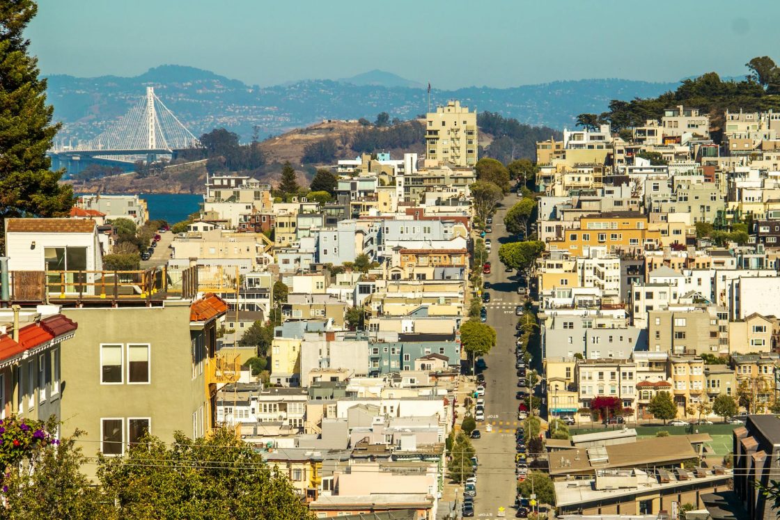 Paisagem urbana de San Francisco, com residências alinhadas em terreno inclinado e ponte ao fundo próxima à baía