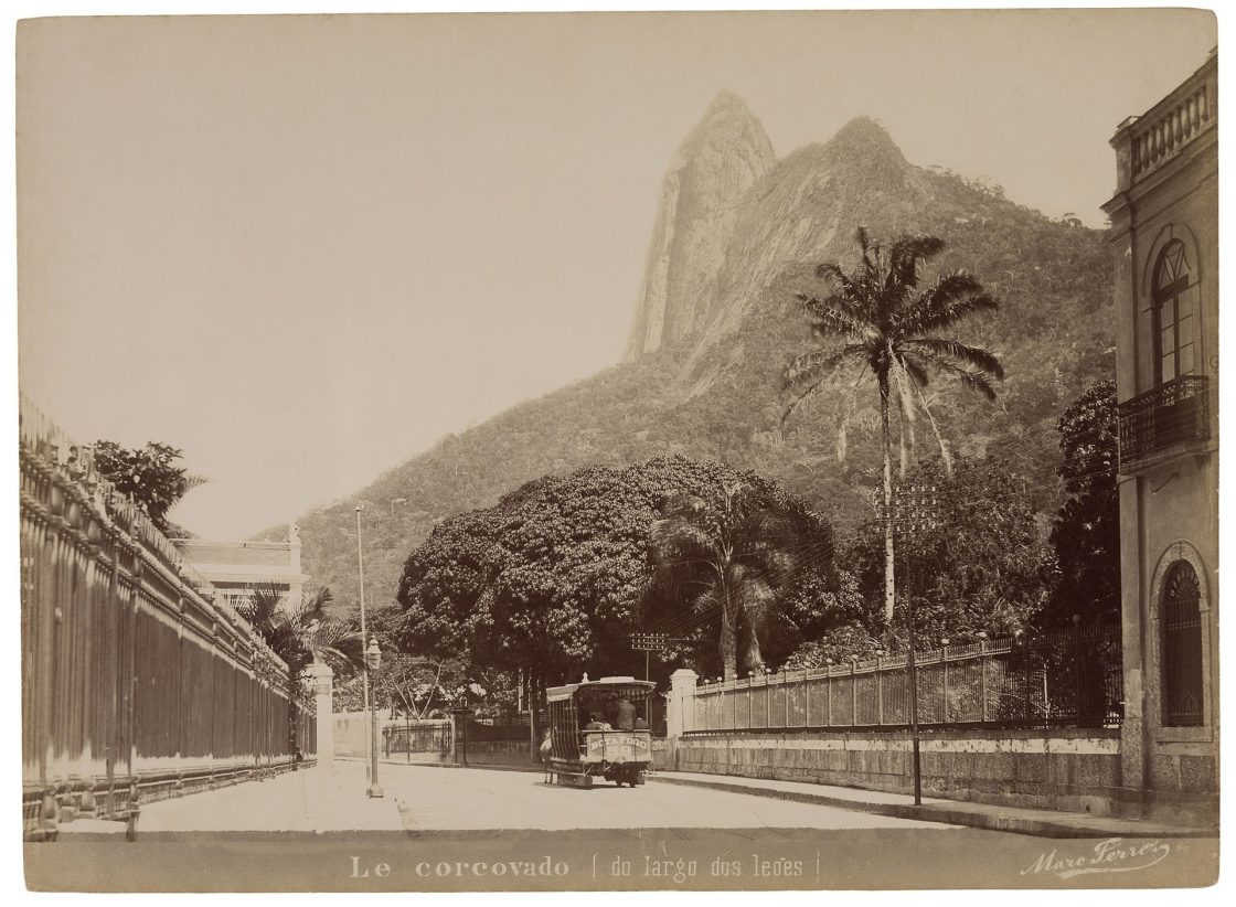 Fotografia do século 19 mostrando o Morro do Corcovado sem o Cristo Redentor, com rua, bonde, palmeiras e construções no primeiro plano e a montanha ao fundo