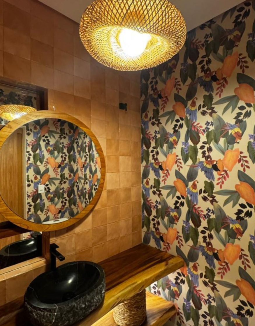 Lavabo com uma parede decorada com papel de parede botânico vibrante, exibindo araras coloridas entre folhas tropicais e frutas alaranjadas.