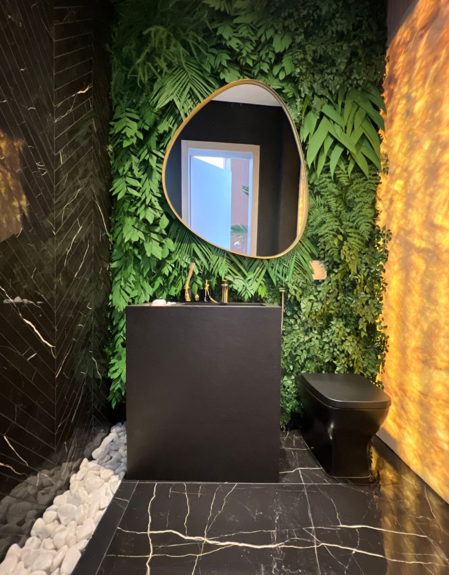 Lavabo com conceito de design biofílico, apresentando parede com jardim vertical e espelho orgânico, bancada preta e painel de pedra ônix retroiluminada