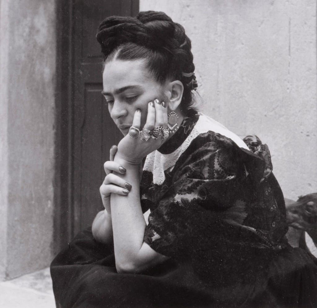 Retrato em preto e branco de Frida Kahlo sentada na Casa Azul, com expressão introspectiva, apoiando o rosto na mão, usando vestimenta tradicional mexicana e acessórios