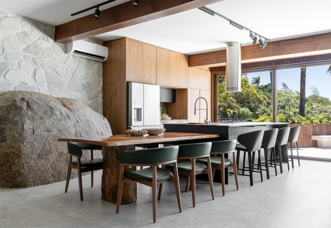 Cozinha com ilha e mesa para refeições, ampla abertura para o exterior e parede com pedras naturais ao fundo