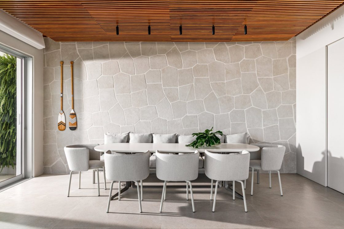 Sala de jantar com mesa ampla para oito lugares e parede de mosaico de revestimento que reproduz pedra off-white ao fundo