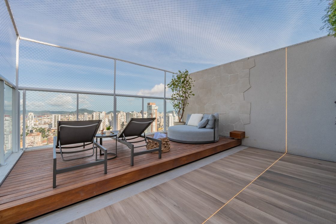 Área externa de cobertura de prédio com piso em porcelanato que reproduz madeira. Também há um deck de madeira com espreguiçadeiras e um sofá. Na pareda do lado direito ainda há um revestimento que reproduz pedra