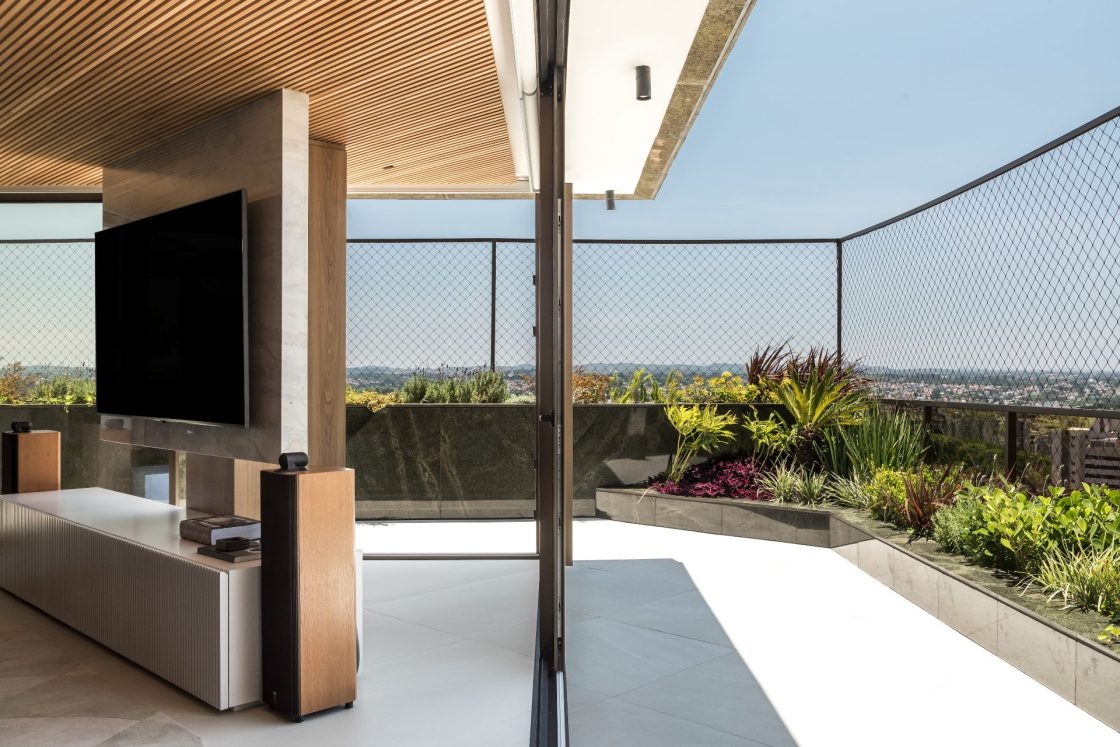 Varanda de apartamento com vista para a cidade e integração com a sala de TV, ambos os ambientes com o mesmo revestimento que reproduz pedra no piso