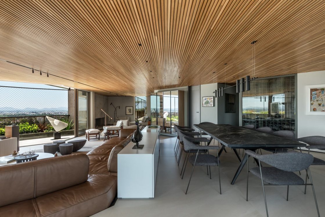 Sala de estar com mesa retangular em tom escuro, integrada ao living com sofá de couro marrom. Ambiente tem teto em madeira ripada