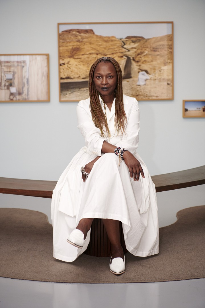 Koyo Kouoh, curadora da Bienal de Veneza 2026, sentada em um branco, em frente a obras de arte penduradas na parede. Ela usa roupa e sapatos brancos