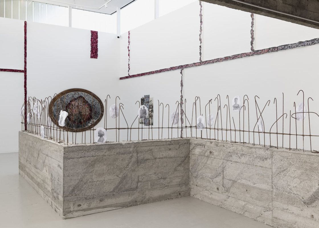 Instalação artística apresentada na Bienal de Veneza combina estruturas de concreto, barras metálicas enferrujadas, fotografias e elementos orgânicos em espaço expositivo minimalista de paredes brancas